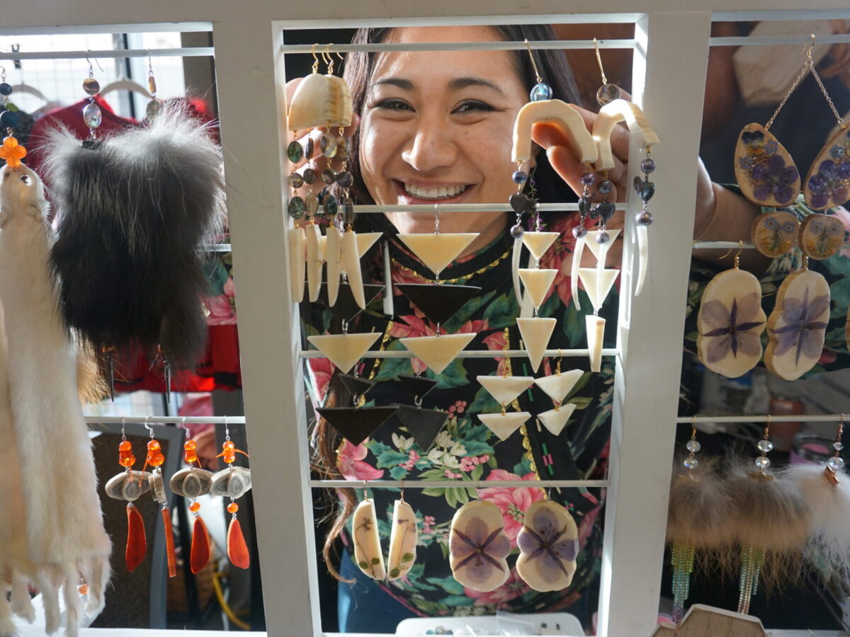 Britt'Nee Brower of Utqiagvik peers through hanging jewelry at her table at the Alaska Federation of Natives convention on Oct. 16, 2025. Brower creates works of art out of a variety of media. Among her skills is carving, sewing, beading, etching, fashion design and poetry. She is among the artists listed in the Alaska Native Arts Directory. (Photo by Yereth Rosen/Alaska Beacon)